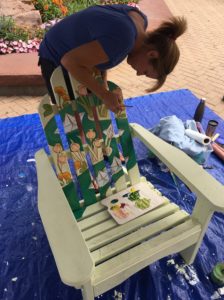 woman painting an Adirondack chair depicting an image of paper boats floating down a river