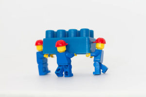 Three LEGO men carrying a large blue LEGO block
