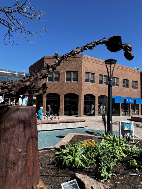 A sculpture of connected hats, made of rusted steel, hats cantalever over a bed of flowers