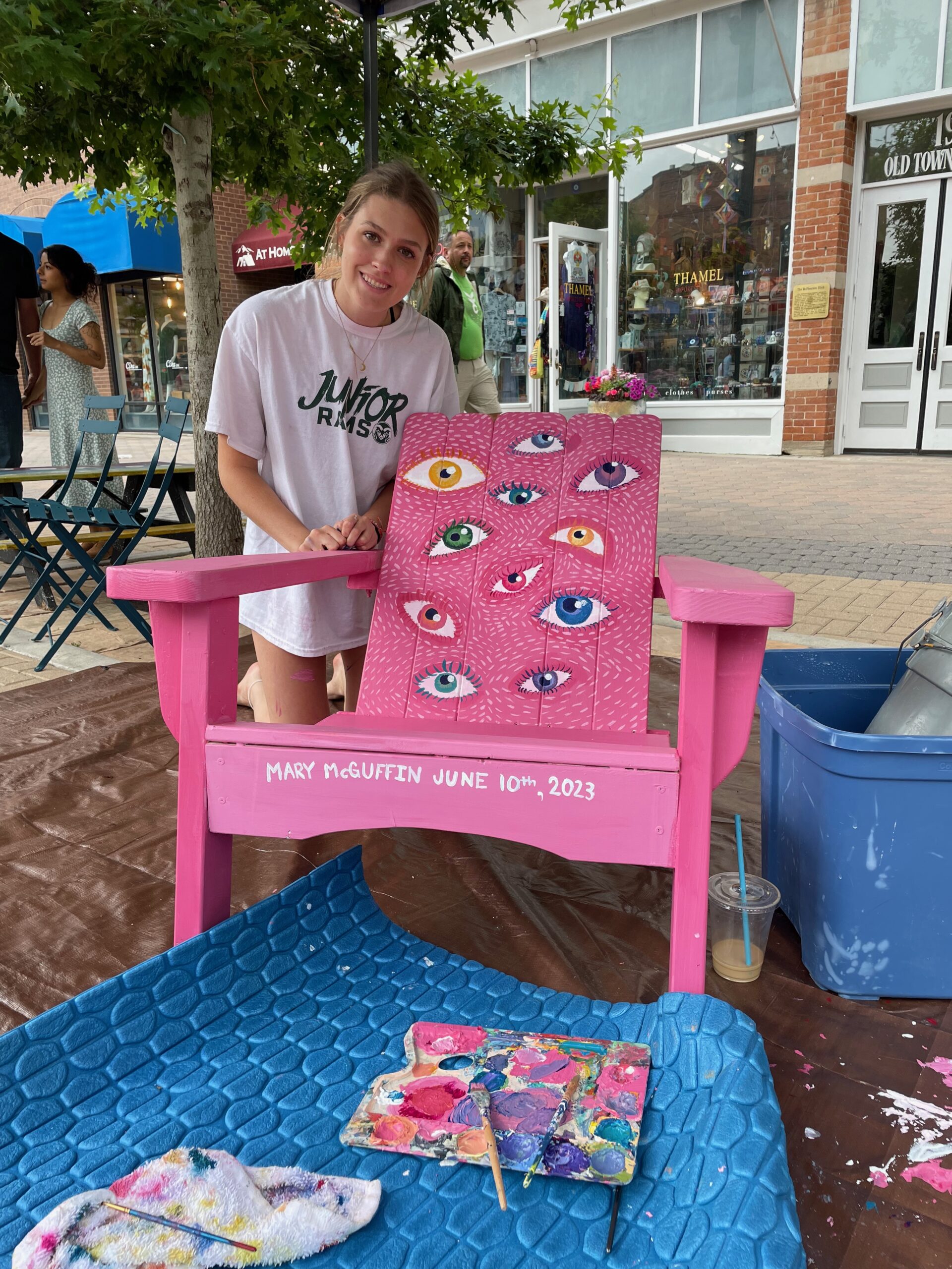 woman displaying painted Adirondack chair with multiple eyeballs painted with pink background